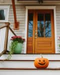 Pumpkin with carved face on porch steps, flanked by green pots with red flowers, leading to a wooden front door.