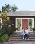 Couple sitting on brick steps of a brown house with a pink door, surrounded by greenery and large windows.