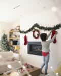 Woman hanging stockings by a fireplace, with a decorated Christmas tree and cozy living room setting.