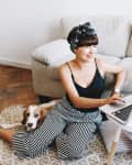 Woman in striped pants using a laptop on the floor, with a dog beside her and a gray sofa in the background.