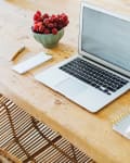 Open laptop on wooden table with a bowl of grapes, notepad, pen, and smartphone on stacked notebooks.