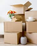 Stacked cardboard boxes with books, a vase, and a cup on a wooden floor near a white sofa.
