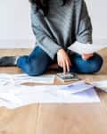 Person sitting on wooden floor, using a calculator, surrounded by scattered bills and documents.