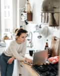 Woman in a cozy kitchen leaning on a counter, using a laptop, with kitchen utensils and plants in the background.