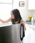 Woman opening a refrigerator in a bright kitchen with white cabinets, yellow flowers, and a cutting board on the counter.