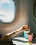 Hand stirring drink in pink cup on airplane tray table next to window.