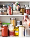 Pantry shelf with jars of grains, pasta, canned goods, and a hand reaching for a jar.