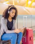 Woman with headphones sitting in airport, using smartphone, next to red suitcase.