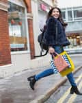 Woman in blue coat and jeans joyfully jumping on a city street, holding colorful shopping bags.