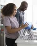 Couple washing dishes in a bright kitchen, smiling and drying a blue container.