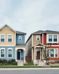 Three colorful suburban houses in a row: yellow, blue, and red, with small front yards and trees.