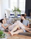 Two women laughing on a beige sofa in a bright living room with a wooden table and potted plant.