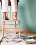 Person standing on a chair painting a wall light blue, with paint can and tray on the floor.