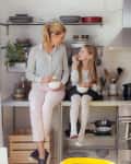 Mother and daughter eating in a modern kitchen with open shelves, fruit bowls, and stainless steel appliances.