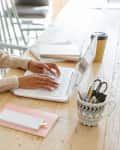Person typing on a laptop at a wooden table with notebooks, a coffee cup, and a pen holder.