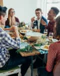 Group of friends laughing and dining together at a rustic wooden table with wine glasses and plates of food.