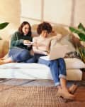 Two people sitting on a white sofa with a laptop, surrounded by potted plants in a cozy room.