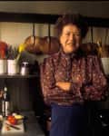 Woman in a patterned blouse standing in a kitchen with hanging copper pots, utensils, and a cutting board with ingredients.