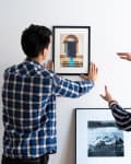 Two people in plaid shirts hanging framed art on a white wall.