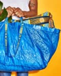 Person holding a large blue shopping bag with a plant and patterned fabric inside against a yellow background.