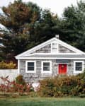 Small cottage with gray shingles, red door, surrounded by lush greenery and tall trees.