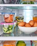 Refrigerator shelves with eggs in a bowl, sliced meats, broccoli, and various fruits in glass containers.