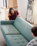 Couple moving a teal sofa in a room with moving boxes, a red chair, and a ladder.