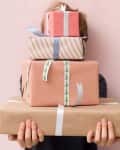 Person holding a stack of wrapped gifts with various ribbons against a pink background.