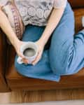 Person sitting on a brown leather sofa, holding a coffee mug, wearing blue jeans and white sneakers.