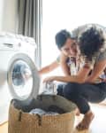 Couple doing laundry together, smiling near a washing machine with detergent bottles on top and a wicker basket on the floor.