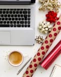 Laptop on desk with red wallet, coffee, pen, gift wrap, bows, and a knitted sweater.