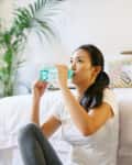 Woman sitting on floor drinking water in a cozy bedroom with plants and patterned pillows.