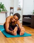 Woman stretching on a blue yoga mat in a living room with plants and a gray sofa.