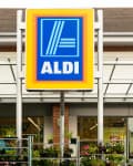 Aldi storefront with large blue and yellow sign, glass windows, and potted plants outside.
