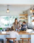 Family in a kitchen with wooden countertops, pendant lights, and open shelves. A child sits on the counter.