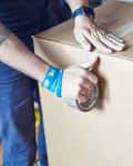 Person sealing a large cardboard box with packing tape, wearing a smartwatch and colorful wristbands.
