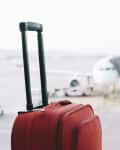 Red suitcase with extended handle in airport terminal, airplane visible through window.