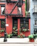 Red brick building with black fire escape, potted plants, and window boxes filled with greenery and flowers.