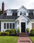 White two-story house with black door, dormer windows, and manicured shrubs in front yard.