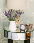 Bedside table with lavender in a white vase, a lamp, a clock, and a decorative box.