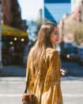 Woman in a yellow floral dress crossing a city street with blurred traffic and buildings in the background.