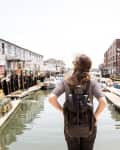Person with backpack standing on a dock, overlooking a canal with boats and industrial buildings.