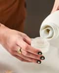 Person pouring liquid detergent into a cap, next to a stack of white towels and a jar of clothespins.