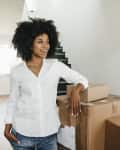 Woman in white shirt leaning on stacked moving boxes in a bright hallway.