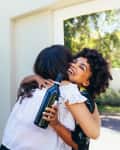 Two women hugging outdoors, one holding a wine bottle, near a leafy bush.