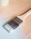 Paintbrush applying light blue paint on wooden surface next to open paint can lid.