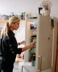 Woman in a kitchen opening a refrigerator, surrounded by shelves with jars, bottles, and kitchen items.