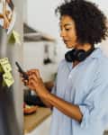 Woman in a kitchen using a smartphone, wearing headphones around her neck, with photos and sticky notes on the fridge.