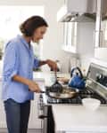 Woman in blue shirt cooking on a stove in a bright kitchen with a blue kettle.