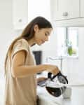 Woman pouring coffee into a mug in a bright kitchen with white cabinets and a knife block.